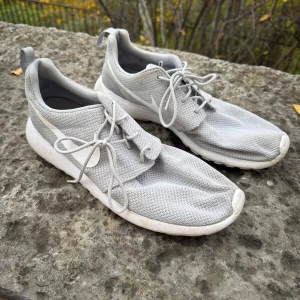 Grå Nike sneakers med mesh - Säljer ett par grå Nike sneakers med vit sula och vit swoosh på sidan. Skorna har rund tå, snörning och är tillverkade i ventilerande meshmaterial som gör dem riktigt sköna. Perfekta för dig som gillar stilrena och sportiga skor.