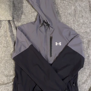 Grå och svart vindjacka Under Armour - Snygg vindjacka från Under Armour i grått och svart med huva och vit logga på bröstet. Jackan har en praktisk bröstficka med dragkedja och elastiska muddar. Perfekt för dig som gillar sportig stil och vill ha något lätt och funktionellt. Knappt använt, skick = helt ny. 