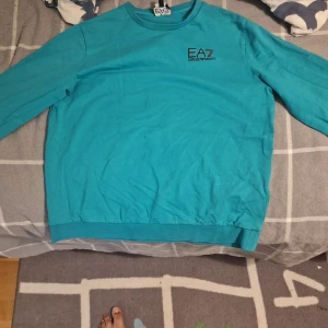 tröja från EA7  - Turkos långärmad tröja från EA7 Emporio Armani med rund hals och diskret logga på bröstet. Tröjan är i mjuk bomull och har ribbade muddar vid ärmslut och nederkant. Perfekt för dig som gillar stilrena och bekväma plagg.