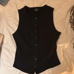 Svart väst med knappar från Gina Tricot - Stilren svart väst från Gina Tricot i storlek M. Västen har en klassisk rak passform och är försedd med svarta knappar längs hela framsidan. Materialet känns mjukt och har en lätt stretch, perfekt för att skapa en clean look. Rund halsringning och enkla linjer.