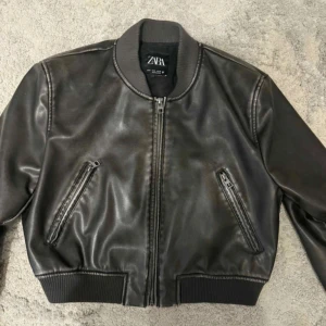 Svart bomberjacka i skinn från Zara - Snygg svart bomberjacka från Zara i skinnimitation med ribbade muddar och krage. Jackan har två fickor med dragkedja och klassisk zip framtill. Perfekt för dig som vill ha en stilren och cool look.