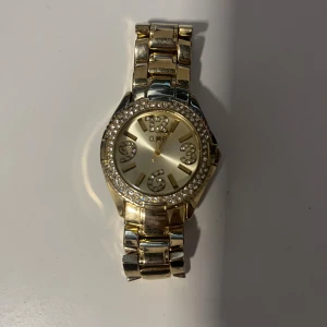 Guldig klocka med glittrande stenar från O.N.E - Snygg guldig klocka från O.N.E med guldarmband och rund urtavla. Urtavlan är dekorerad med glittrande stenar runt kanten och siffrorna 3, 6, 9 och 12 är markerade med glittriga stenar. Klockan har en klassisk och stilren look som sticker ut.