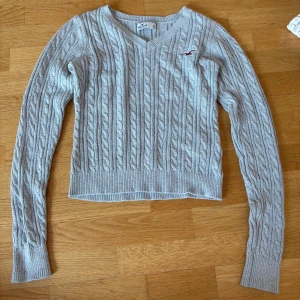 Ljusgrå kabelstickad tröja Hollister - Supersnygg ljusgrå kabelstickad tröja från Hollister med v-ringning och lång ärm. Klassisk logga broderad på bröstet 💕