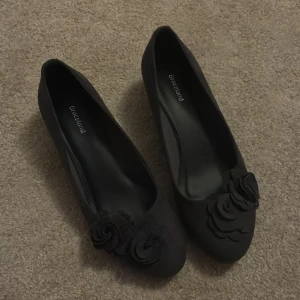 Svarta pumps med blommor Graceland - Snygga svarta pumps från Graceland med rund tå och dekorativa blommor framtill. Skorna har en låg klack och är tillverkade i mocka-liknande material som ger en mjuk känsla. Perfekt för dig som gillar stilrena detaljer och vill ha något extra på fötterna.