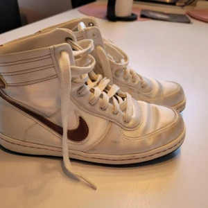 Nike Air Force 1 High vit/brun - Säljer ett par Nike Air Force 1 High sneakers i vit med bruna detaljer och brunt swoosh. Klassisk high-top modell med snörning och rund tå. Ovandel i syntet och skinn, slitstark gummisula med Air-dämpning. Perfekt för dig som gillar retrostil och streetwear.