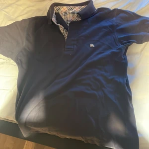 Blå Burberry kortärmad skjorta - Snygg mörkblå kortärmad skjorta från Burberry med klassiskt rutigt mönster på insidan av kragen. Skjortan har broderad logga på bröstet och knappar framtill. Perfekt för dig som gillar stilrena och ikoniska detaljer.