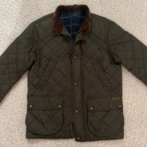 Quiltad grön jacka från Polo Ralph Lauren - Quiltad mörkgrön jacka från Polo Ralph Lauren med bruna manchesterdetaljer på kragen. Jackan har flera fickor med tryckknappar, dragkedja och snygga rutiga detaljer på insidan.  NYPRIS: Ca. 5000 kr