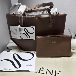 Brun handväska Polène Cyme Mini - Snygg brun handväska från Polène, modell Cyme Mini, med stilren och kantig form. Väskan är tillverkad i strukturerat skinn och har matchande clutch med guldfärgad dragkedja. Perfekt storlek för dina essentials och har vita kontrastsömmar.