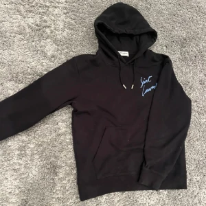 Svart hoodie från Saint Laurent - Svart hoodie från Saint Laurent med vit broderad logga på bröstet. Tröjan har huva med snörning och en stor magficka. Tillverkad i mjuk bomull och har långa ärmar. Perfekt för dig som gillar stilrena designer och exklusiva märken.