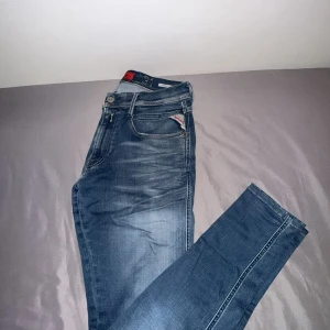 Blå Replay Anbass jeans herr - Säljer ett par blå Replay Anbass jeans med snyggt slitna detaljer och klassisk femficksdesign. Jeansen har normal passform och smalare ben, med Replay-logga på fickan och läderpatch bak i midjan. Perfekta för dig som gillar stilrena och bekväma jeans.