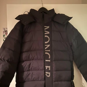 Svart Moncler pufferjacka med huva - Svart pufferjacka från Moncler med stor logotyp längs dragkedjan framtill. Jackan har en rejäl huva, quiltad design och långärmade ärmar. Materialet är syntet och dun, perfekt för kalla dagar. Snygg och modern look med reflekterande detaljer på loggan.