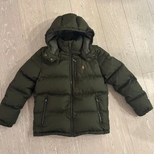 Mörkgrön vinterjacka från Polo Ralph Lauren Lauren - Grön vinterjacka med avtagbar huva från Polo Ralph Lauren. Jackan har dragkedja framtill och två fickor med dragkedja på sidorna. Åt det mindre hållet storleksmässigt. Jättefin jacka som är i princip som ny, mycket sparsamt använd. Bild 5 är lånad från Kids Brand store. 