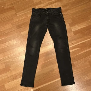 Svarta Replay Anbass jeans, stl 34 - Säljer ett par svarta Replay Anbass jeans i storlek 34. Jeansen har en slim passform med klassisk femficksdesign och snygga slitningar för en cool look. Materialet är stretchigt och bekvämt, perfekt för dig som gillar stilrena svarta jeans.