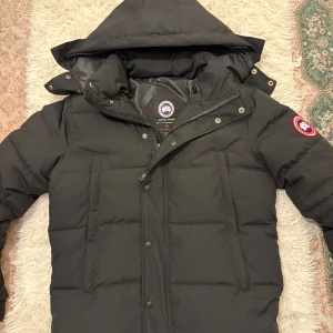 Svart dunjacka från Canada Goose - Svart dunjacka från Canada Goose med huva och klassisk logga på ärmen. Jackan har quiltad design, dragkedja och knappar framtill samt två stora fickor. Perfekt för kalla vinterdagar och har en clean, stilren look. Storlek XS men lite för stor, så passar både S/M, päls medföljer, samt Canada goose prislapp! 