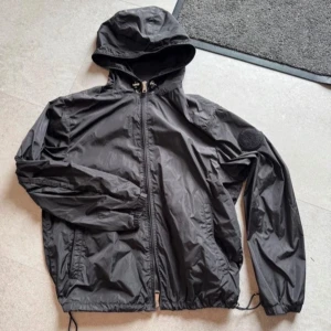 Moncler windbreaker - Svart vindjacka från Moncler med huva och dragkedja framtill. Jackan har Moncler-logga på ärmen, justerbar nederkant med snörning och är tillverkad i lätt polyester. Perfekt för dig som gillar stilrena och funktionella plagg. Inga defekter använd ca 3 gånger. Skicket bedöms vara nytt. Kom privat för mer frågor! Frakt inom 24H