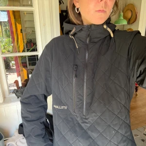 Svart quiltad jacka Stellar Equipment - Svart quiltad jacka från Stellar Equipment med huva och halv dragkedja framtill. Jackan har ett diskret mönster, bröstficka med dragkedja och justerbara snören i huvan. Perfekt för kyliga dagar och snygg till streetwear.