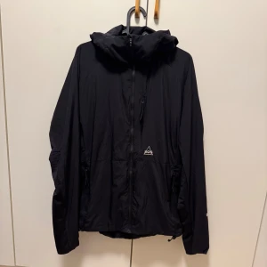 Svart vindjacka från SOS, storlek S - Svart vindjacka från SOS med huva och hel dragkedja framtill. Jackan har minimalistisk design, diskret logga på bröstet och praktiska fickor med dragkedja. Perfekt för blåsiga dagar och enkel att matcha med din streetwear.