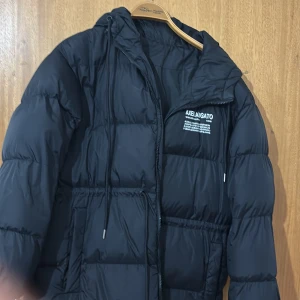 Svart pufferjacka Axel Arigato - Svart pufferjacka från Axel Arigato med huva och snörning. Jackan har en stilren design med logotyptryck på bröstet, dragkedja framtill och långa ärmar. Materialet är syntet och jackan är perfekt för kalla dagar. Passformen är normal och modellen är klassisk.