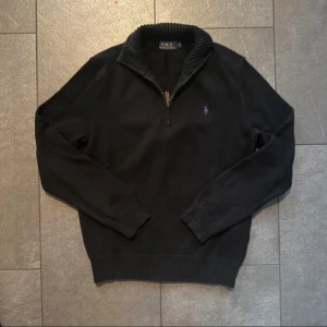 Svart stickad tröja Polo Ralph Lauren - Svart stickad tröja från Polo Ralph Lauren i storlek S. Tröjan har en ribbad krage, half zip-dragkedja och lila broderad logga på bröstet. Långärmad med ribbade muddar och klassisk passform. Perfekt för dig som gillar stilrena och tidlösa plagg.