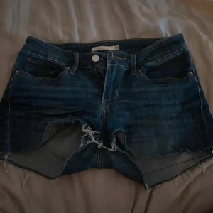 Levi's jeansshorts med rå kant - Säljer ett par klassiska blå jeansshorts från Levi's med råa, fransiga kanter och femficksmodell. Shortsen har normal passform och är tillverkade i bomull. Perfekta för sommaren och har den ikoniska Levi's-lappen bak.