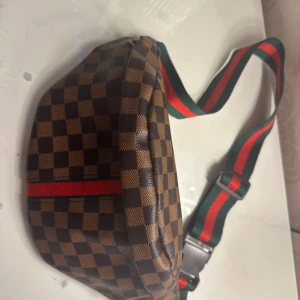 Brun rutig magväska med röd/grön rem - Snygg magväska i brunt och svart rutigt mönster med justerbar rem i grönt och rött. Väskan har dragkedja och är tillverkad i ett syntetmaterial med slät yta. Perfekt för att bära det viktigaste när du är på språng.