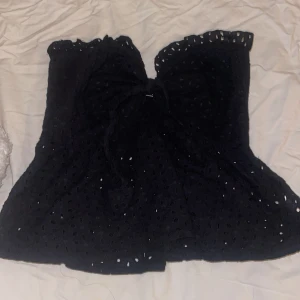 Svart spetsig tubtopp från SHEIN - Svart tubtopp från SHEIN med broderade spetsdetaljer och volangkant upptill. Toppen är axelbandslös och har en söt knytning framtill. Materialet är lätt och luftigt, perfekt för varma dagar. Passformen är normal och toppen har ett romantiskt uttryck.