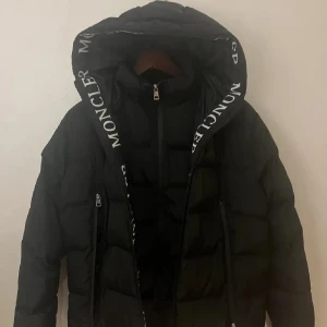 Svart Moncler pufferjacka med huva - Säljer en svart pufferjacka från Moncler med stor huva och tydlig logga längs kanten. Jackan har två dragkedjeförsedda fickor, quiltad design och är långärmad. Perfekt för dig som vill ha en snygg och varm jacka med streetkänsla.