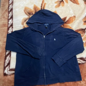 Marinblå hoodie från Polo Ralph Lauren - Snygg marinblå hoodie från Polo Ralph Lauren med dragkedja och klassisk logga broderad på bröstet. Tröjan har huva, långa ärmar och kängurufickor. Tillverkad i mjuk bomull, perfekt för chill dagar.