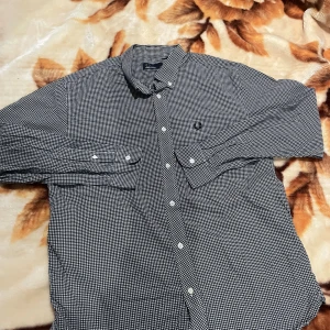 Svartvit rutig Fred Perry skjorta L - Snygg svartvit rutig skjorta från Fred Perry i storlek L. Skjortan har klassisk krage, vita knappar och lång ärm. Broderad Fred Perry-logga på bröstet. Tillverkad i 100% bomull för skön känsla och bra passform.