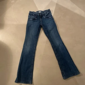 Blå bootcut jeans med justerbar midja - Snygga blå jeans med bootcut passform och klassisk femficksdesign. Jeansen har en justerbar midja med resår och knapp på insidan, vilket gör dem extra bekväma. Materialet är slitstarkt denim och färgen är mörkblå med lätt tvättade detaljer.