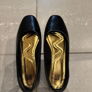 Svarta ballerinaskor med guldinnersula - Stilrena svarta ballerinaskor från XIT med fyrkantig tå och platt sula. Insidan har en glansig guldinnersula med vågigt mönster som ger extra edge. Yttermaterialet är slätt skinnliknande syntet och designen är klassisk men modern.