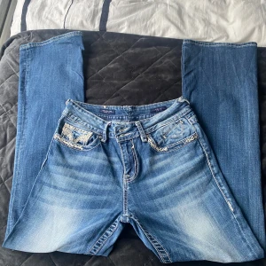 Blå bootcut jeans med broderade fickor Viggos - Snygga blå bootcut jeans från The New York i storlek 10. Jeansen har broderade bakfickor med detaljerade mönster och kontrastsömmar. Klassisk femficksmodell med dragkedja och knapp. Materialet är jeans med en tvättad look och ljusare partier framtill.