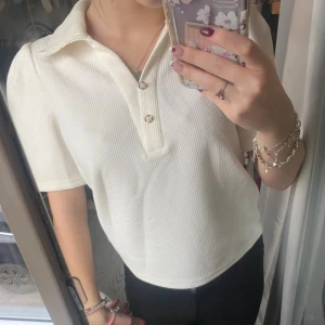 Vit ribbad polotopp från Dazy - Säljer en vit polotopp från Dazy i ribbad struktur med korta ärmar och två snygga guldfärgade knappar vid halsen. Toppen har en klassisk krage och är i ett mjukt material. Perfekt för dig som gillar stilrena och enkla plagg.