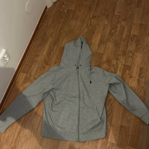 Grå hoodie från Ralph Lauren - Säljer en stilren grå hoodie från Ralph Lauren med dragkedja och klassisk huva. Helt oanvänd eftersom den va för stor, fick den på min födelsedag. Inga hål eller defekter, den har inga tecken på användning heller. Pris kan diskuteras.