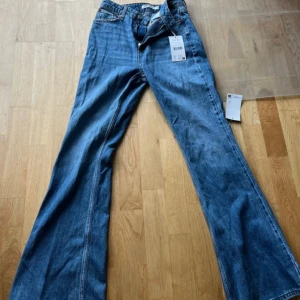 Blå bootcut jeans från NA-KD, strl 36 - Snygga blå jeans från NA-KD i storlek 36 med klassisk bootcut passform. Jeansen har hög midja, dragkedja och knapp framtill. Tillverkade i jeansmaterial med en cool, lite retro vibe. Perfekta för dig som gillar en avslappnad men trendig stil.