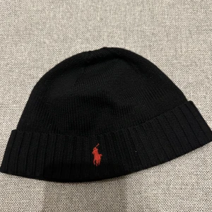 Polo Ralph Lauren mössa - Säljer en svart mössa från Polo Ralph Lauren i mjuk merinoull med ribbad kant och klassisk röd Polo-logga broderad framtill. Mössan är i toppskick, nypris ca 800kr.