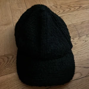 Svart teddykeps från H&M Divided - Svart keps från H&M Divided i one size, tillverkad i mjukt teddy-material som ger en fluffig och cool look. Kepsen har klassisk form med böjd skärm och justerbar rem bak. Perfekt för dig som vill ha en chill och trendig accessoar.