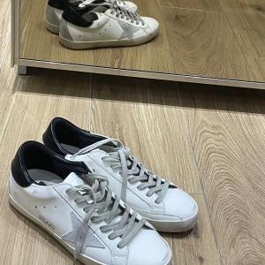 Golden Goose vita sneakers med stjärna - Golden Goose sneakers i vitt skinn med grå mockastjärna på sidan och grå snörning. Svart hälparti och platt sula. Snygg och stilren design med coola detaljer som metalltext på sidan. Perfekt för dig som gillar streetstyle och vill sticka ut. Litet hål i båda hälarna på skorna.