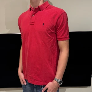 Röd Ralph Lauren piké  - Säljer en klassisk röd Polo t-shirt från Ralph Lauren i storlek M. T-shirten har korta ärmar, krage med knappar och den ikoniska broderade Polo-loggan på bröstet. Tillverkad i 100% bomull för skön och luftig känsla. Perfekt för dig som gillar stilren och tidlös design.