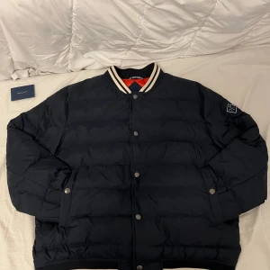 Svart pufferjacka från GANT XL - Snygg svart pufferjacka från GANT i storlek XL med röd insida och coola vita ränder på kragen. Jackan har tryckknappar, dragkedja och ribbade muddar. Perfekt för kalla dagar och har en sportig vibe med klassisk bomberstil. Skriv gärna vid frågor eller föer bilder!