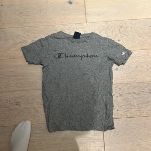 Grå Champion t-shirt med logga - Gråmelerad t-shirt från Champion i storlek Large, med klassisk Champion-logga tryckt över bröstet och en liten logotyp på ärmen. T-shirten har rund hals och korta ärmar, tillverkad i mjuk bomull som är skön mot huden.