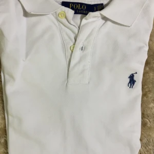 Vit pikétröja från Polo Ralph Lauren - Klassisk vit pikétröja från Polo Ralph Lauren med blå broderad logga på bröstet. Tröjan har krage, knäppning med två knappar och är tillverkad i mjuk bomull. Perfekt för en clean och stilren look.