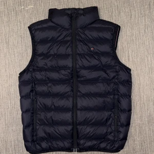 Mörkblå dunväst från Tommy Hilfiger - Snygg mörkblå dunväst från Tommy Hilfiger med quiltad design och diskret logga på bröstet. Västen har dragkedja framtill och två sidofickor med dragkedja. Perfekt för lager-på-lager och en clean look.