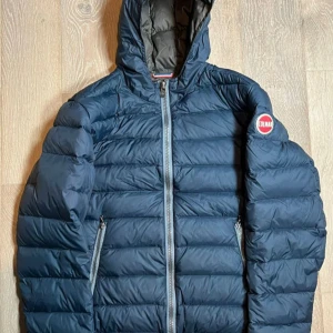 Mörkblå dunjacka från Colmar - Snygg mörkblå dunjacka från Colmar med huva och två dragkedjeförsedda fickor. Detta är en förbeställing snabbast frakt  3 dagar. Jackan har quiltad design och en tydlig logga på ärmen. Perfekt för kyliga dagar och har en sportig vibe. Storlek M/48.