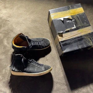 Saint Laurent svarta höga sneakers mocka - Svarta höga sneakers från Saint Laurent i mocka med fransdetaljer längs skaftet och snörning framtill. Insidan är fodrad med lyxigt gult skinn och yttersulan är vit med Saint Laurent Paris-logga. Snygg och unik design som sticker ut.