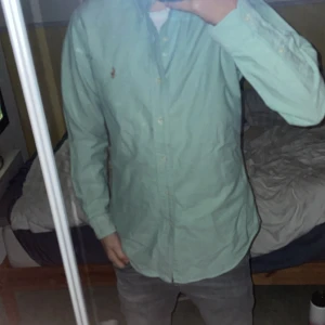 Grön skjorta från Ralph Lauren - Snygg grön skjorta från Ralph Lauren med klassisk button-down krage och broderad logga på bröstet. Skjortan har lång ärm, knappar framtill och är gjord i mjuk bomull. Perfekt för dig som gillar stilrena och tidlösa plagg.