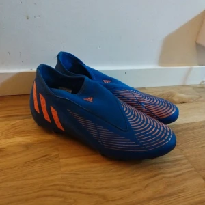 Blå-orange adidas fotbollsskor - Säljer ett par blå fotbollsskor från adidas med orangea detaljer och mönster. Skorna har en strömlinjeformad design utan snörning och är tillverkade i syntetmaterial. Perfekta för dig som vill sticka ut på planen med både stil och funktion.