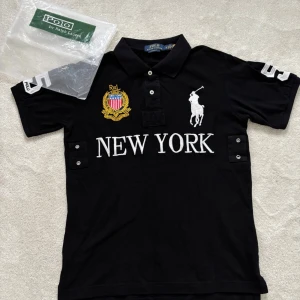Svart pikétröja Polo Ralph Lauren S - Svart pikétröja från Polo Ralph Lauren med broderad New York-text, stort vitt polospelare-märke och guldfärgad emblem på bröstet. Nummer 5 i vitt på ryggen och ärmen. Klassisk krage, kortärmad och tillverkad i bomull. Custom fit-modell med snygga knappar framtill.