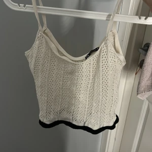 Vit stickad linnetopp från Zara - Supersöt vit stickad linnetopp från Zara med smala axelband och svart kant nertill. Toppen har ett virkat mönster som ger en chill och somrig vibe. Perfekt att styla med jeans eller kjol för en trendig look.