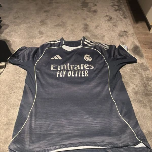 Real Madrid Mbappé fotbollströja blå - Säljer en mörkblå Real Madrid fotbollströja från Adidas med Mbappé och nummer 9 på ryggen. Tröjan har korta ärmar, vita detaljer och tryck med 'Emirates Fly Better' på bröstet. Materialet är lätt och andas, perfekt för träning eller match.